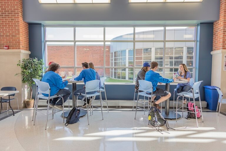 Seven students sitting at two tables in a cafeteria, with a large window next to them letting in sunlight