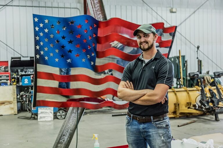 Man in workshop standing in front of steel replica of the American flag